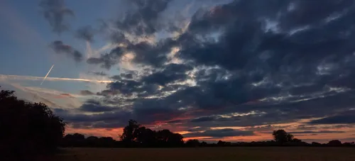 This landscape photograph captures a late sunset against a copse of trees, with dramatic clouds filling the sky. Taken in the late evening during the summer, the image highlights the glow of the setting sun as it casts vibrant hues of orange and red along the horizon. The agricultural fields in the foreground emphasize the rural nature of the scene, while the silhouettes of the trees provide a natural focal point. The presence of clouds adds texture to the sky, enhancing the overall atmosphere of the sunset and contributing to the tranquil yet striking summer evening in the countryside.